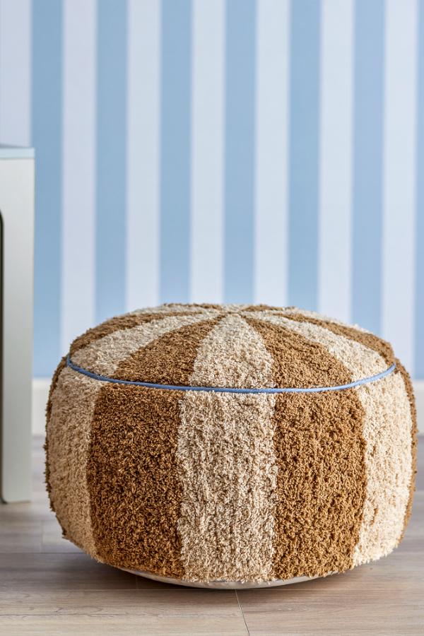 Charoline Pouf Brown – Bloomingville MINI: Pouf vor einer Wand mit blau gestreifter Tapete.