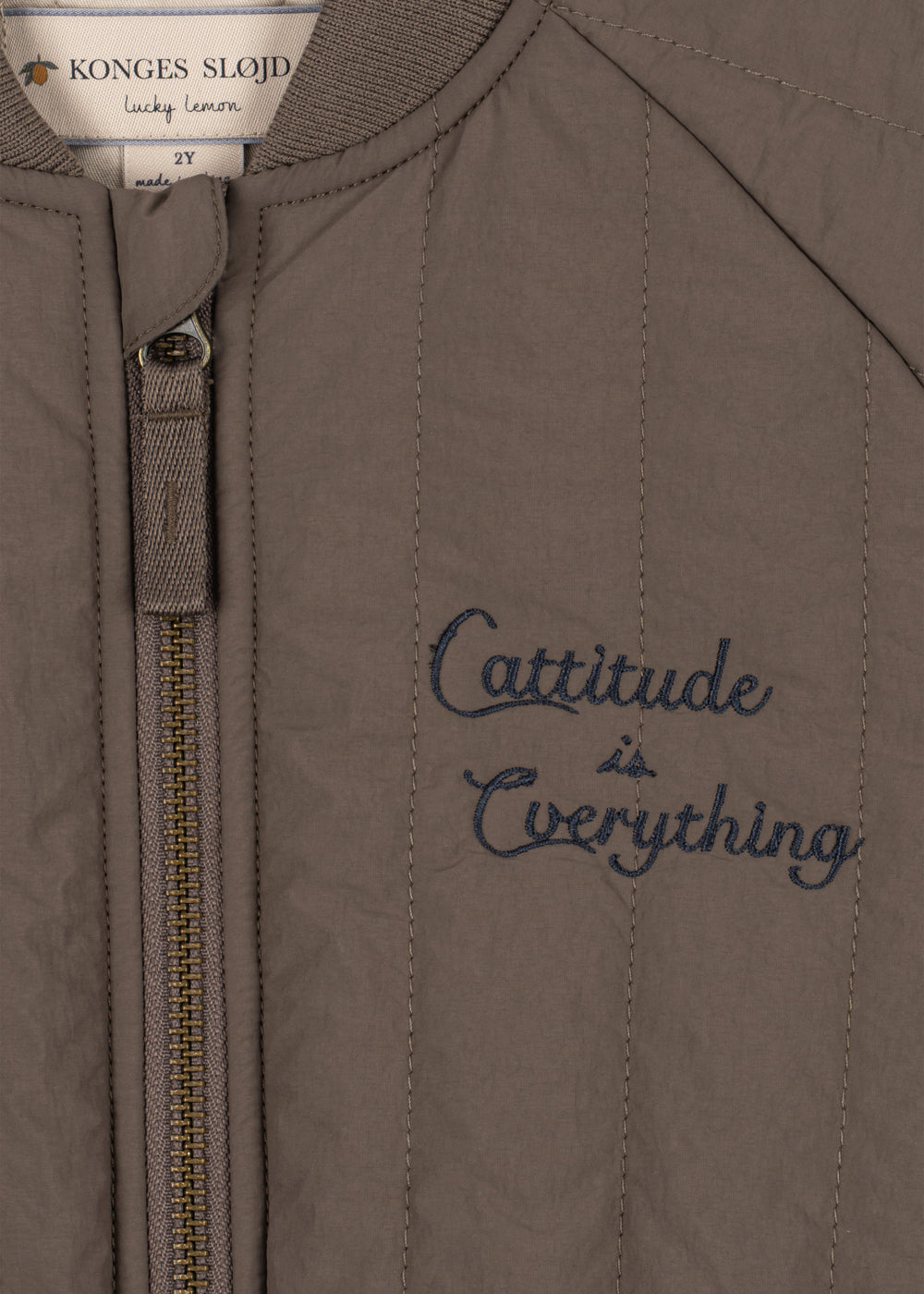 Veste thermique Juno Bomber – Walnut – Konges Sløjd : détail de la fermeture éclair et du texte brodé sur la veste marron.