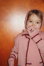 Woll-Balaclava Collette – Pale Blush – Konges Sløjd: Kind mit rosa Kirschen-Strickjacke hält den Ärmel vors Gesicht.